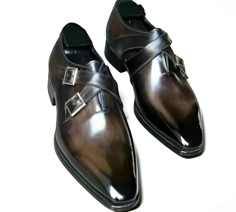 Op de afbeelding: Een paar bruin lederen geklede schoenen met een glanzende afwerking. De schoenen hebben een dubbel monk strap design met zilveren gespen en een spitse neus. De schoenen worden op een witte achtergrond gepresenteerd.
