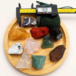 May include: A collection of nine raw, unpolished mineral stones in various colors including yellow, red, black, white, pink, green, and clear, arranged on a round bamboo tray. A black digital caliper displays a measurement of 46.8 mm.