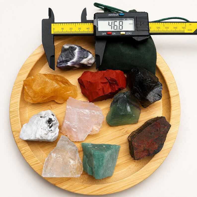 May include: A collection of nine raw, unpolished mineral stones in various colors including yellow, red, black, white, pink, green, and clear, arranged on a round bamboo tray. A black digital caliper displays a measurement of 46.8 mm.