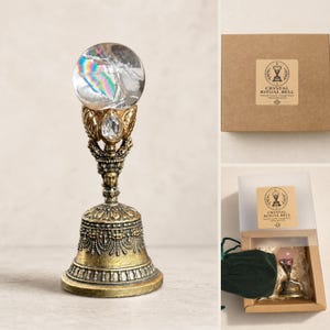 May include: A ritual bell featuring a clear crystal sphere top with iridescent reflections, set upon an ornate, antique-gold finished metal base with a small teardrop crystal accent. The bell is packaged in a brown box labeled Crystal Ritual Bell.