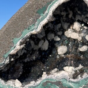 May include: A geode featuring a dark gray, textured outer shell with a vibrant green mineral band bordering the interior cavity. The hollow center is filled with small, translucent white crystalline structures that catch the light.
