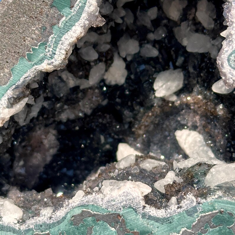 May include: A geode featuring a deep, dark, and reflective interior cavity lined with numerous small, translucent white crystalline structures. The outer rim of the geode displays a textured, layered surface with shades of dark green and gray.
