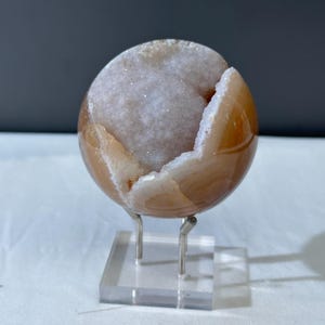 May include: A spherical, polished agate geode, showcasing a warm, amber-toned exterior and a sparkling, crystalline interior. The geode is mounted on a clear acrylic stand with silver-colored metal supports, creating a floating effect. The geode is a decorative item.