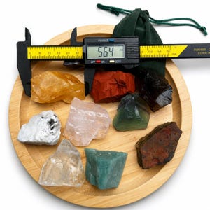 May include: A collection of nine raw, multi-colored mineral stones arranged on a round bamboo tray. A black digital caliper measures one stone at 56.4 mm. The stones include shades of yellow, red, black, white, pink, green, and brown.