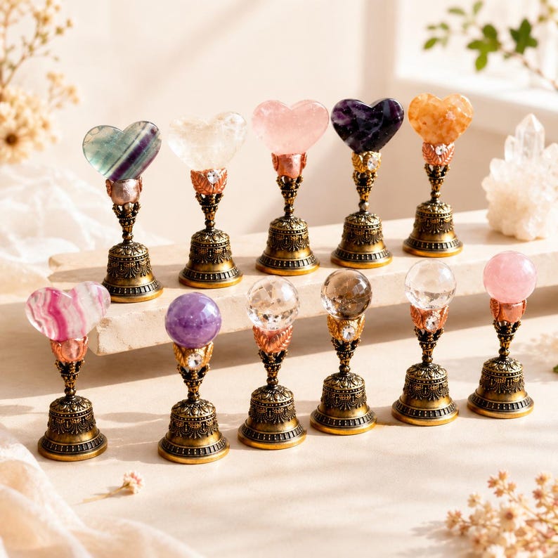 May include: Eleven decorative ornaments featuring polished gemstone tops in heart and sphere shapes, mounted on ornate, antique-style brass-toned metal bell bases with intricate filigree detailing and small embedded clear crystals.