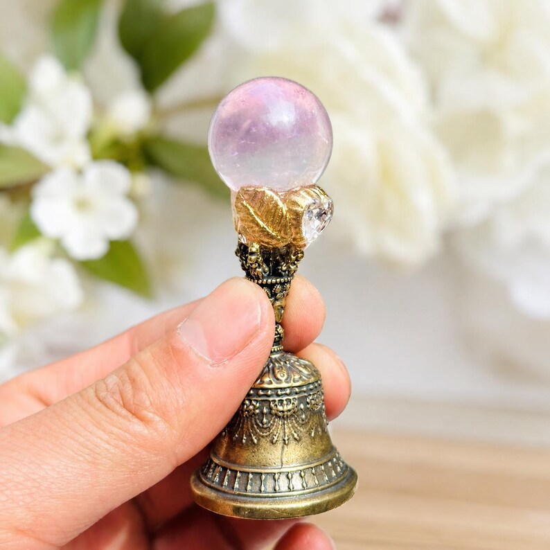 May include: A small decorative object featuring a translucent pale pink sphere resting atop a metallic gold-toned base. The base has an ornate, bell-shaped design with intricate filigree patterns and a textured leaf-like accent near the top.