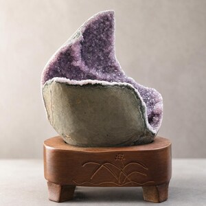 May include: A large amethyst geode, showcasing a vibrant purple crystal interior, is displayed on a small, carved wooden stand. The geode's exterior has a rough, gray-green appearance, contrasting with the sparkling crystals within.