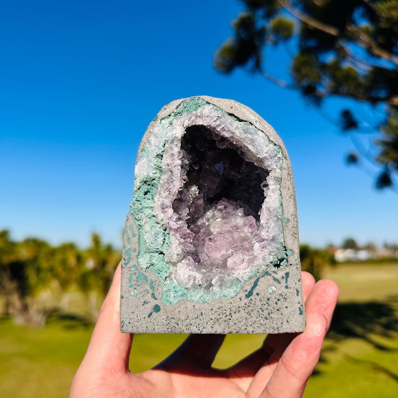 May include: A dome-shaped amethyst geode featuring a hollow center filled with light purple, crystalline structures. The exterior is composed of a smooth, grey, speckled stone casing with a distinct green mineral border surrounding the crystals.