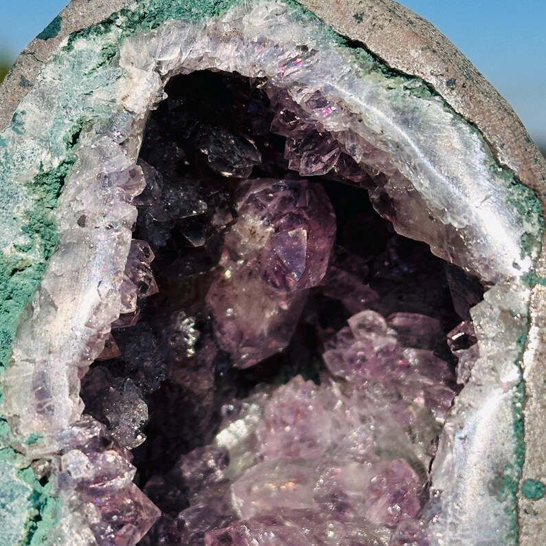 May include: A geode featuring a hollow center filled with translucent purple amethyst crystals. The outer rim of the rock formation is a rough, textured gray stone with a distinct layer of vibrant green mineral deposits lining the inner edge.