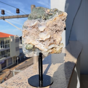 May include: A natural stone specimen in shades of white, beige, green, and yellow, mounted on a black metal display stand. The rough-hewn stone displays a variety of textures and crystalline structures, suitable for decorative purposes.