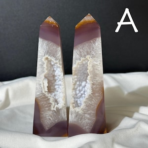 Può includere: Due obelischi di agata lucida. Gli obelischi sono un mix di viola, bianco e marrone. L'interno di ogni obelisco ha una cavità simile a una geode piena di formazioni di cristallo bianco. Gli obelischi sono esposti su un panno bianco.