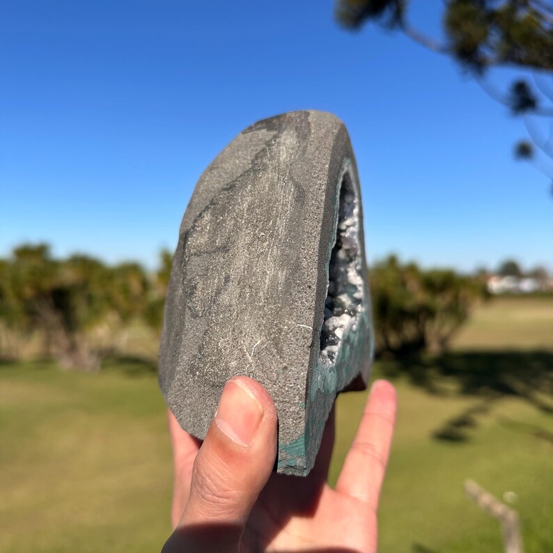 May include: A gray, semi-circular geode with a rough, textured exterior surface. The interior cavity reveals a cluster of dark, crystalline structures. The object is held against a bright blue sky and a blurred green landscape background.