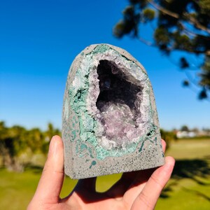 May include: A dome-shaped geode featuring a rough, gray exterior and a hollow center filled with translucent, light purple amethyst crystals. The inner rim of the geode is lined with a textured, pale green mineral layer surrounding the crystals.