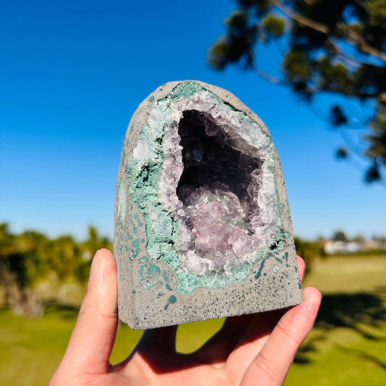 May include: A dome-shaped geode featuring a rough, gray exterior and a hollow center filled with translucent, light purple amethyst crystals. The inner rim of the geode is lined with a textured, pale green mineral layer surrounding the crystals.