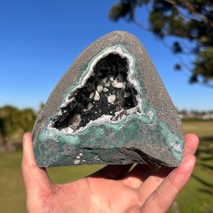 May include: A triangular geode rock featuring a dark gray exterior with patches of green mineral deposits. The center cavity is lined with small, sparkling white and translucent crystals, creating a sharp contrast against the dark, rough stone surface.