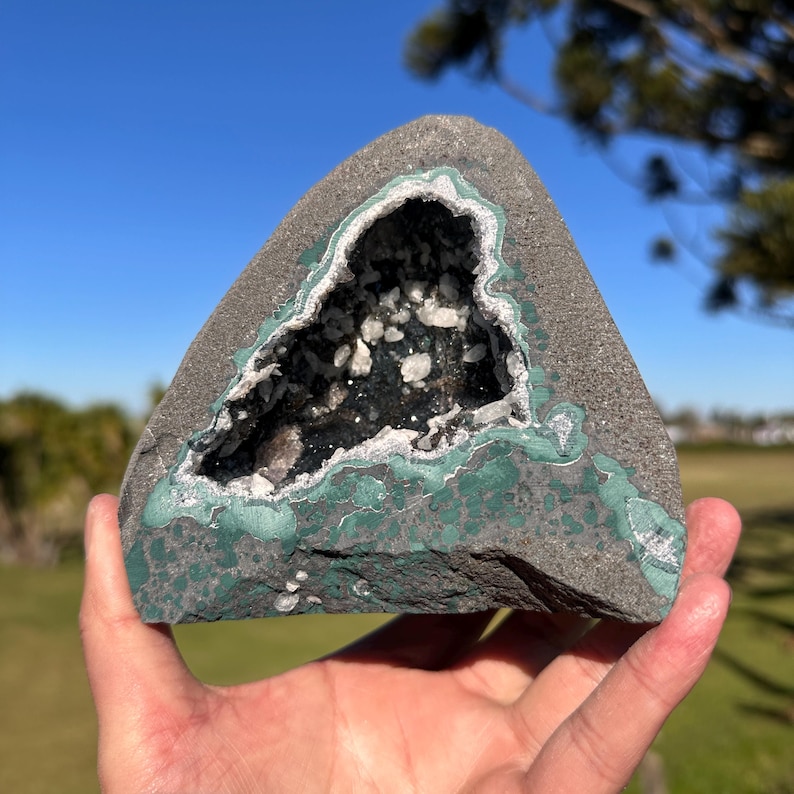 May include: A triangular geode rock featuring a dark gray exterior with patches of green mineral deposits. The center cavity is lined with small, sparkling white and translucent crystals, creating a sharp contrast against the dark, rough stone surface.