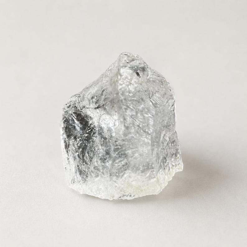 May include: A raw, clear, and translucent crystal stone with an irregular, jagged shape. The surface features numerous sharp facets and natural ridges that catch the light, creating a textured appearance with internal inclusions and cloudy white areas.