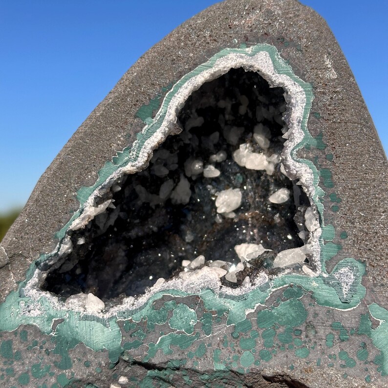 May include: A dark gray geode rock featuring a central cavity lined with small, translucent white crystalline structures. The exterior of the stone displays patches of vibrant green mineral deposits, creating a contrast against the dark rock surface.