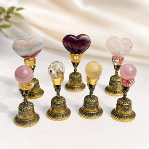 May include: A collection of seven decorative stands, each featuring an antique gold-toned metal bell base. The stands are topped with various polished gemstones, including heart-shaped and spherical stones in shades of pink, purple, and yellow.