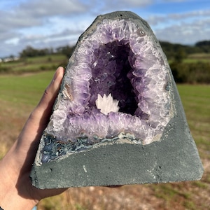 Puede incluir: Una geoda de amatista triangular que presenta un interior cristalino de color púrpura intenso con una prominente formación de calcita blanca en el centro. El exterior está recubierto por una matriz de roca gris oscuro lisa y base plana.