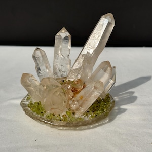 Op de afbeelding: Een cluster van heldere kwartskristallen, sommige met een lichte amberkleur, gerangschikt op een cirkelvormige basis. De basis is versierd met kleine groene en witte stenen, wat een natuurlijke, aardse uitstraling creëert. De kristallen variëren in grootte en vorm.