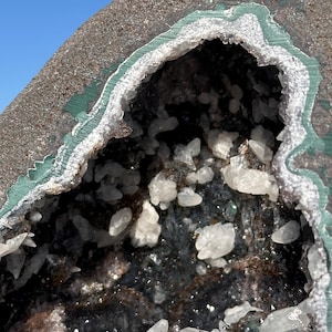 May include: A geode featuring a dark, rough exterior with a green-tinted rim surrounding a hollow center filled with small, translucent white crystalline structures and dark, textured mineral deposits against a clear blue sky background.