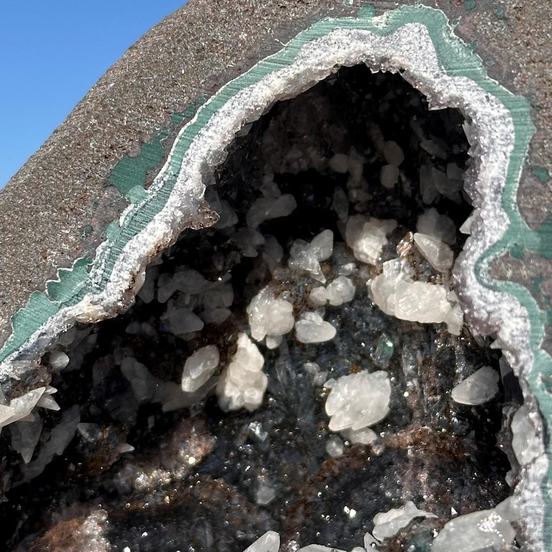 May include: A geode featuring a dark, rough exterior with a green-tinted rim surrounding a hollow center filled with small, translucent white crystalline structures and dark, textured mineral deposits against a clear blue sky background.
