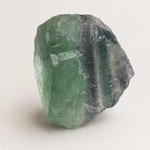 May include: A raw, translucent fluorite crystal specimen featuring a prominent blend of light green and deep purple color zones. The stone has an irregular, angular shape with natural facets, sharp edges, and a smooth, glassy texture throughout.