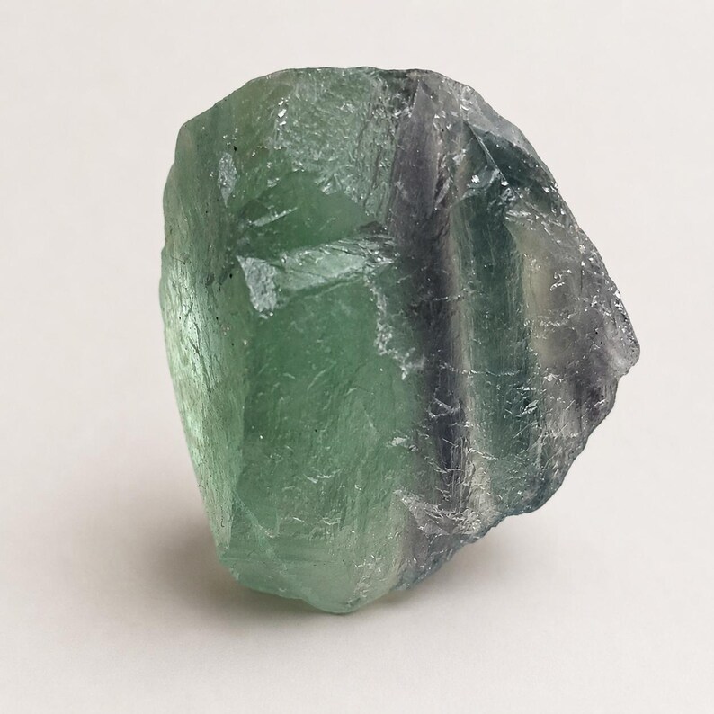 May include: A raw, translucent fluorite crystal specimen featuring a prominent blend of light green and deep purple color zones. The stone has an irregular, angular shape with natural facets, sharp edges, and a smooth, glassy texture throughout.
