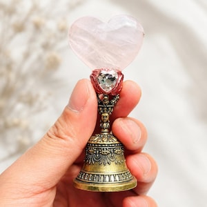 May include: A decorative bell featuring a pale pink heart-shaped stone at the top, accented by a small clear faceted heart-shaped crystal set in a red metal base. The handle is crafted from antique brass-toned metal with intricate relief details.