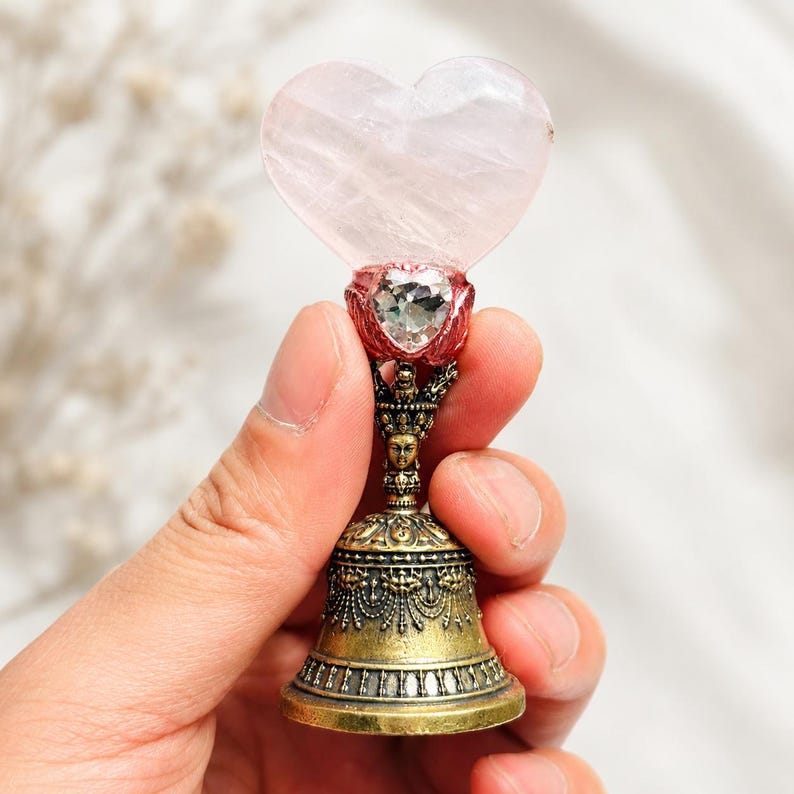 May include: A decorative bell featuring a pale pink heart-shaped stone at the top, accented by a small clear faceted heart-shaped crystal set in a red metal base. The handle is crafted from antique brass-toned metal with intricate relief details.