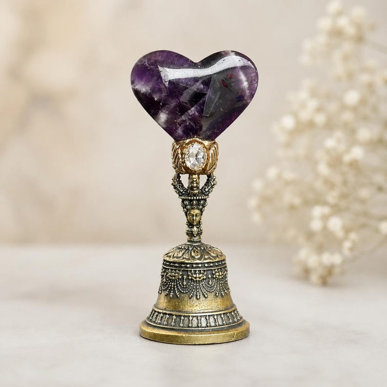 May include: A decorative bell featuring a polished purple heart-shaped amethyst stone at the top. The handle is crafted from antique-toned brass with intricate filigree details, a small clear crystal accent, and a sculpted face at the center.