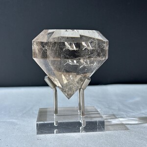 Op de afbeelding: Een helder, facetgeslepen kristal in diamantvorm, gemonteerd op een heldere acrylbasis met zilveren metalen steunen. Het kristal heeft interne insluitsels en staat tegen een donkere achtergrond. Het geheel is een weergave van natuurlijke schoonheid en geometrische precisie.