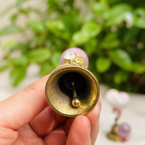 May include: A brass bell with a smooth interior and a central clapper. The handle features a light pink spherical bead topped with a gold-toned decorative element. The bell has a metallic finish and a circular opening, showing the internal structure.