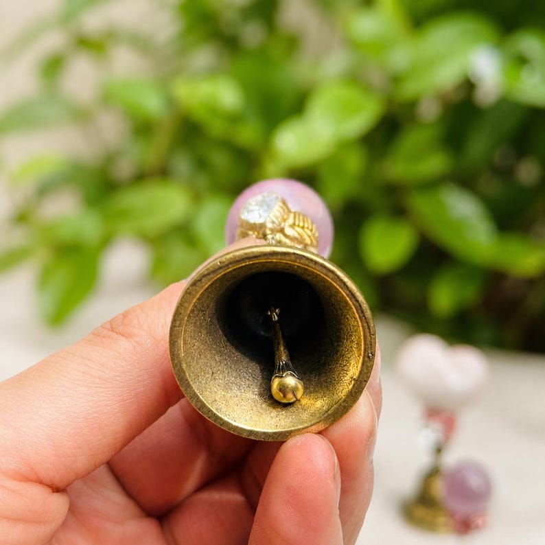 May include: A brass bell with a smooth interior and a central clapper. The handle features a light pink spherical bead topped with a gold-toned decorative element. The bell has a metallic finish and a circular opening, showing the internal structure.