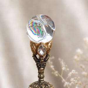 May include: A clear spherical crystal orb with internal rainbow refractions sits atop a metallic gold-toned stand. The stand features an intricate design with a central face, leaf-like accents, and a small, clear, teardrop-shaped faceted stone.