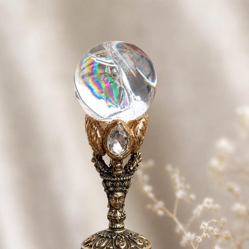 May include: A clear spherical crystal orb with internal rainbow refractions sits atop a metallic gold-toned stand. The stand features an intricate design with a central face, leaf-like accents, and a small, clear, teardrop-shaped faceted stone.
