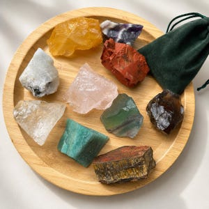May include: A collection of nine raw, unpolished mineral stones in various colors including yellow, purple, red, white, pink, green, brown, and teal, arranged on a round bamboo tray alongside a small, dark green velvet drawstring pouch.