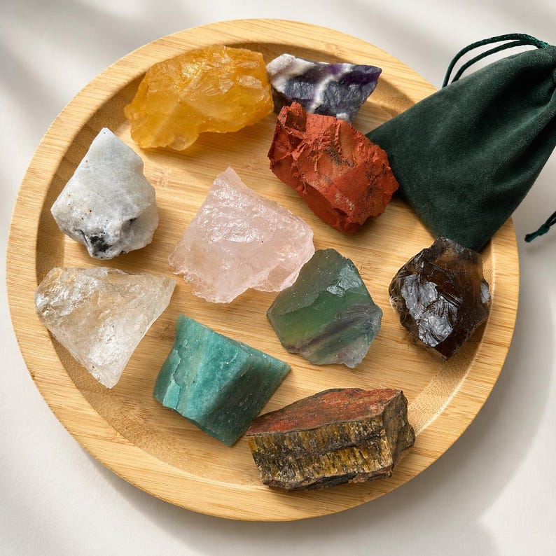 May include: A collection of nine raw, unpolished mineral stones in various colors including yellow, purple, red, white, pink, green, brown, and teal, arranged on a round bamboo tray alongside a small, dark green velvet drawstring pouch.