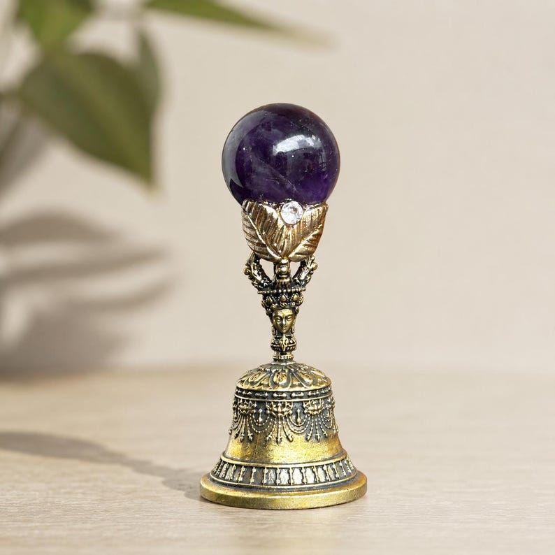 May include: A decorative bell featuring a polished purple amethyst sphere at the top, held by a gold-toned metal mount with leaf-like details and a small clear crystal accent. The handle includes a sculpted face, and the base has intricate patterns.