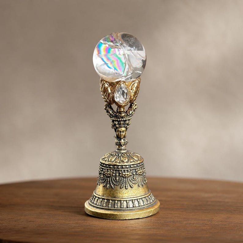 May include: A decorative bell featuring an antique gold-toned metal base with intricate filigree and beaded detailing. The handle is topped with a clear, spherical glass orb that displays internal rainbow light refractions and a teardrop crystal.