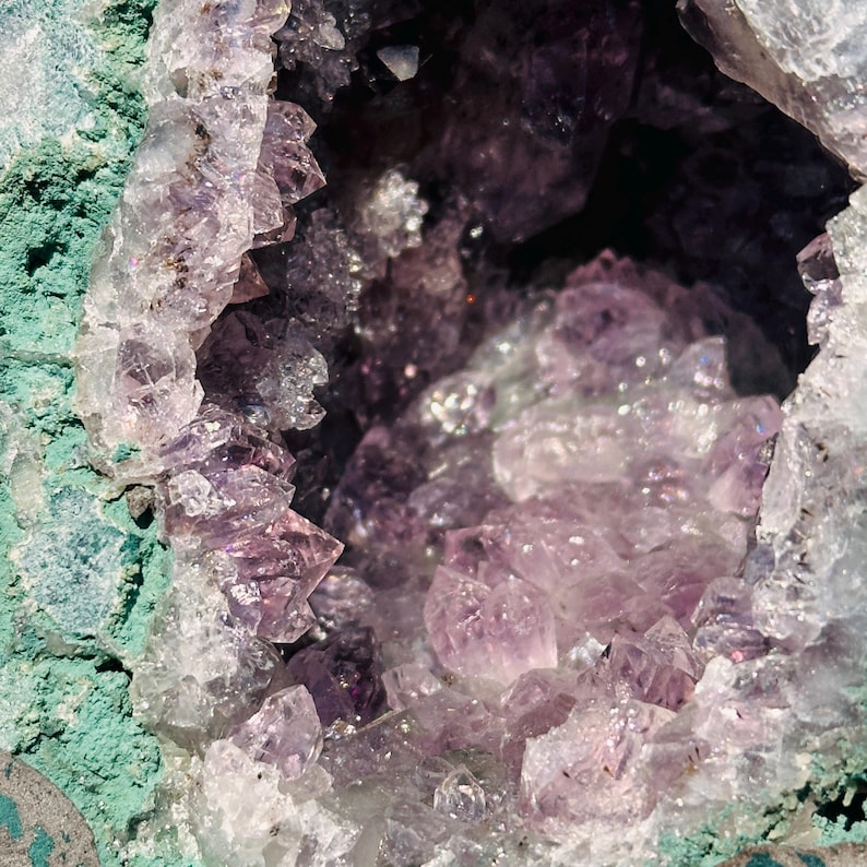 May include: A natural amethyst geode featuring a cluster of translucent, light purple, pointed quartz crystals housed within a rough, textured, teal-colored rock exterior. The interior cavity is filled with jagged, crystalline formations.