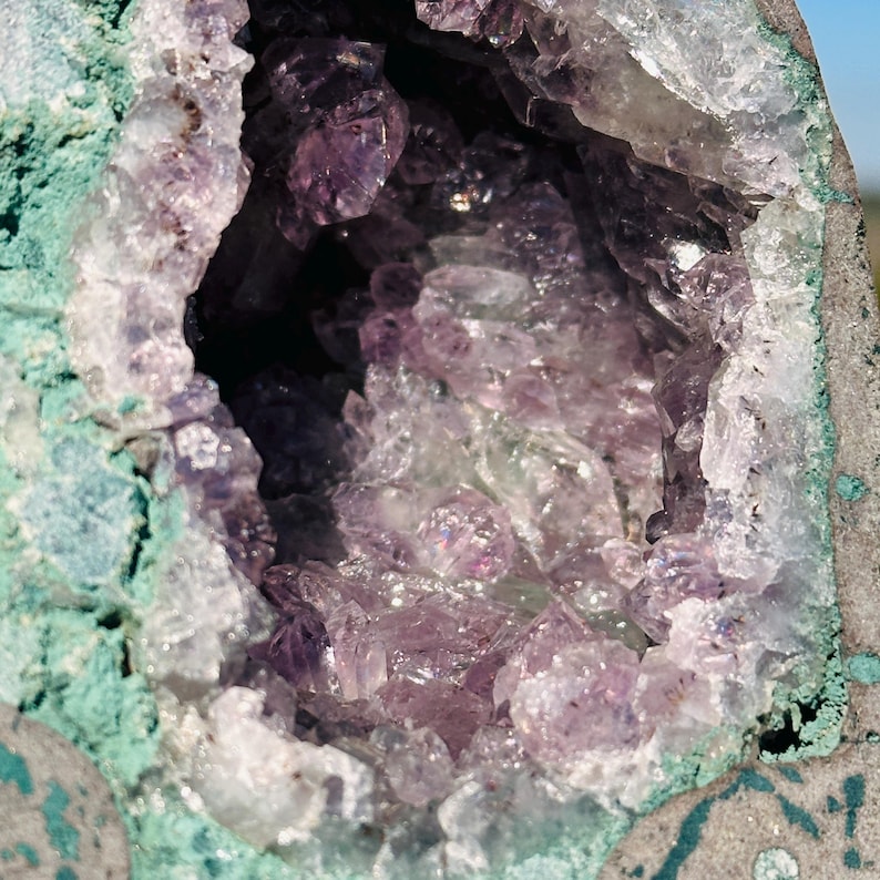 May include: A geode featuring a hollow center filled with translucent, light purple amethyst crystals. The exterior of the rock formation is a rough, textured surface with a distinct teal-green coloration surrounding the crystal cavity.