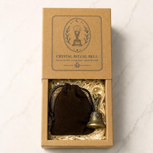 May include: A brown cardboard box containing a dark brown drawstring pouch and a vintage brass bell topped with a clear crystal sphere. The box lid features a black line drawing of a bell and the text CRYSTAL RITUAL BELL Natural Crystal Vintage Brass Handcrafted Gift.