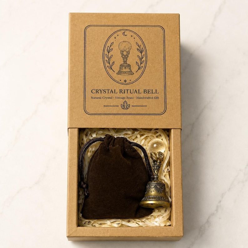 May include: A brown cardboard box containing a dark brown drawstring pouch and a vintage brass bell topped with a clear crystal sphere. The box lid features a black line drawing of a bell and the text CRYSTAL RITUAL BELL Natural Crystal Vintage Brass Handcrafted Gift.