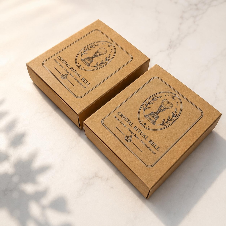 May include: Two square brown cardboard boxes featuring black ink illustrations of a bell topped with a crystal. The text reads CRYSTAL RITUAL BELL, Natural Crystal, Vintage Brass, Handcrafted Gift, with a small crystal icon below the text.