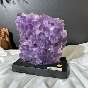Könnte beinhalten: Ein großer, natürlicher Amethyst-Kristallcluster, der einen tiefvioletten Farbton aufweist. Die Kristallformation ist auf einem schwarzen rechteckigen Sockel montiert. Die Oberfläche des Amethysts besteht aus zahlreichen kleinen, kristallinen Strukturen, die ein strukturiertes Aussehen erzeugen. Der Hintergrund ist eine Mischung aus Schwarz und Weiß.