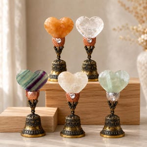 May include: Five decorative ornaments featuring heart-shaped gemstones atop ornate, antique-style brass bell bases. Each piece includes a small, clear crystal accent nestled between the stone heart and the detailed metal stand, showcasing varied colors.