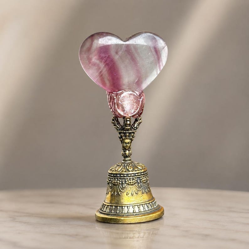 May include: A decorative bell featuring a polished, heart-shaped purple and white fluorite stone at the top. The stone is held by a metallic claw setting atop a detailed, antique-gold colored metal handle and a flared, ornate bell base.