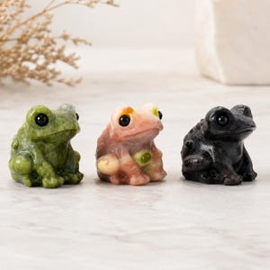 Könnte beinhalten: Drei kleine geschnitzte Froschfiguren aus Stein, die in einer Reihe angeordnet sind. Der erste Frosch ist olivgrün gesprenkelt, der zweite ist blassrosa mit cremefarbenen und grünen Einschlüssen und der dritte ist tiefschwarz. Jeder Frosch hat eine glatte Haut und schwarze Augen.